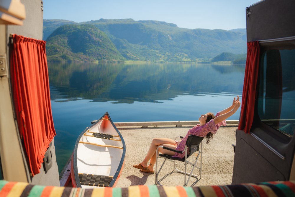 En kvinne slapper av i en sammenleggbar stol ved siden av en campingvogn, strekker armene lykkelig mot solen. Foran henne er en kano plassert ved bredden av et rolig fjord omgitt av frodige fjell.